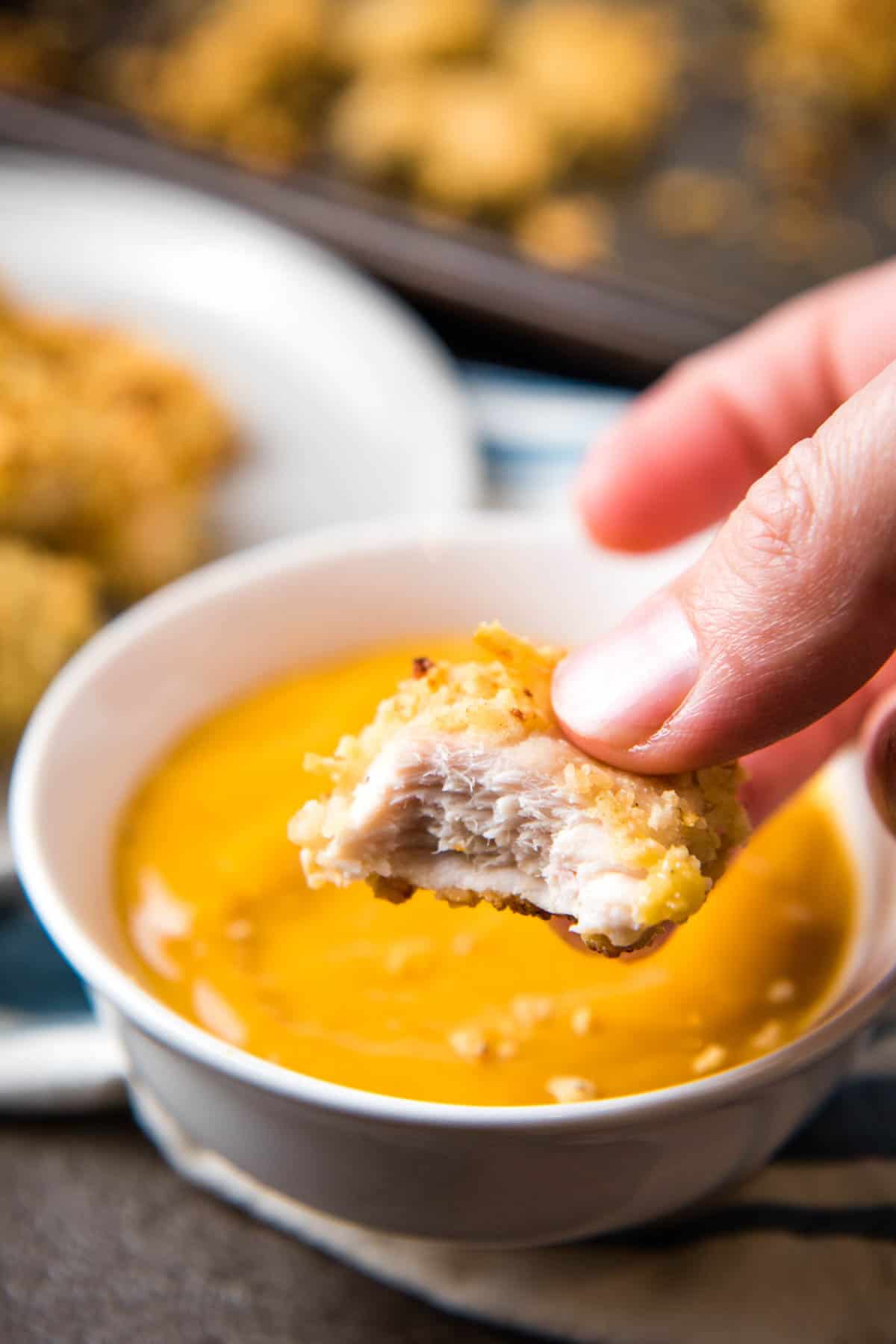 Chicken nuggets on cookie sheet fingers holding chicken Pringler with bite taken out over white bowl with chicken dipping sauce