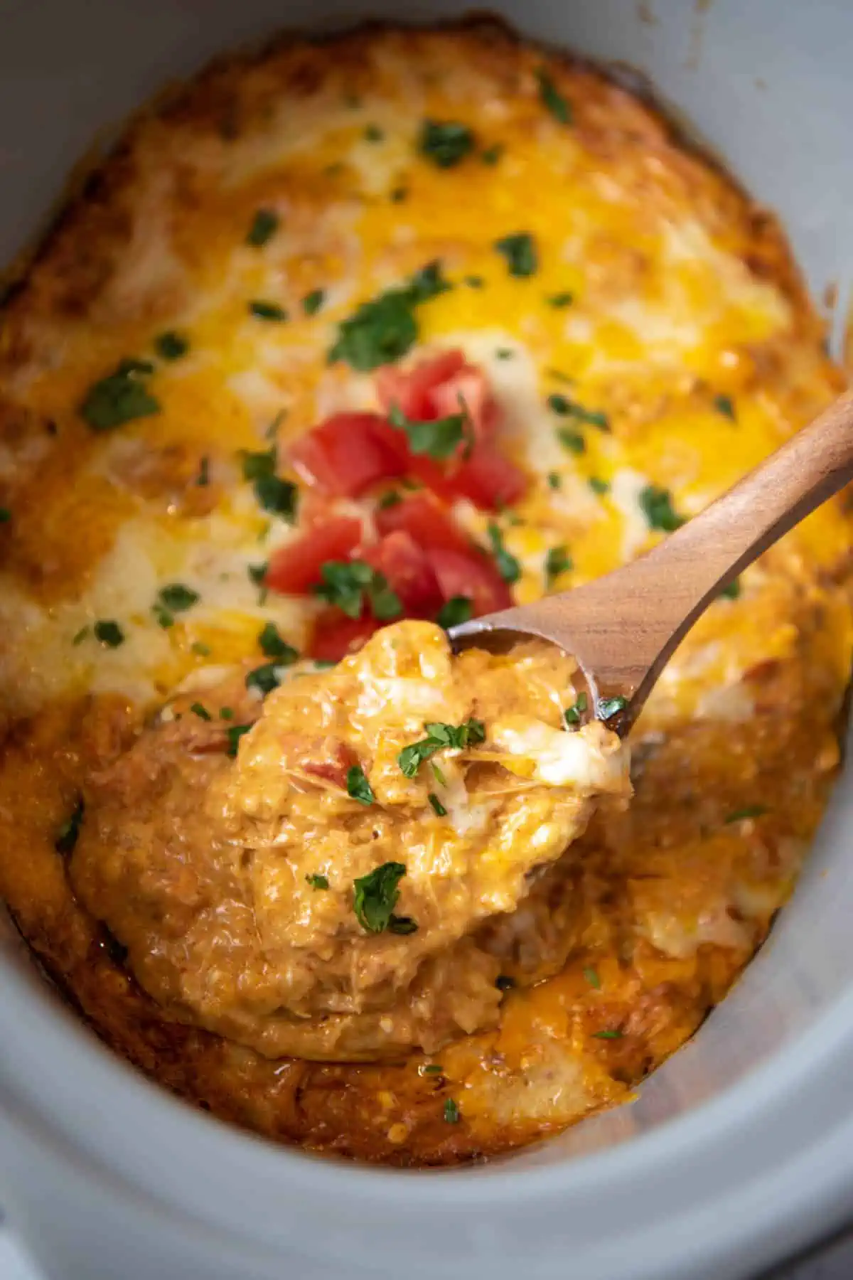 wooden spoonful of CrockPot chicken enchilada casserole over gray slow cooker full of cheesy casserole
