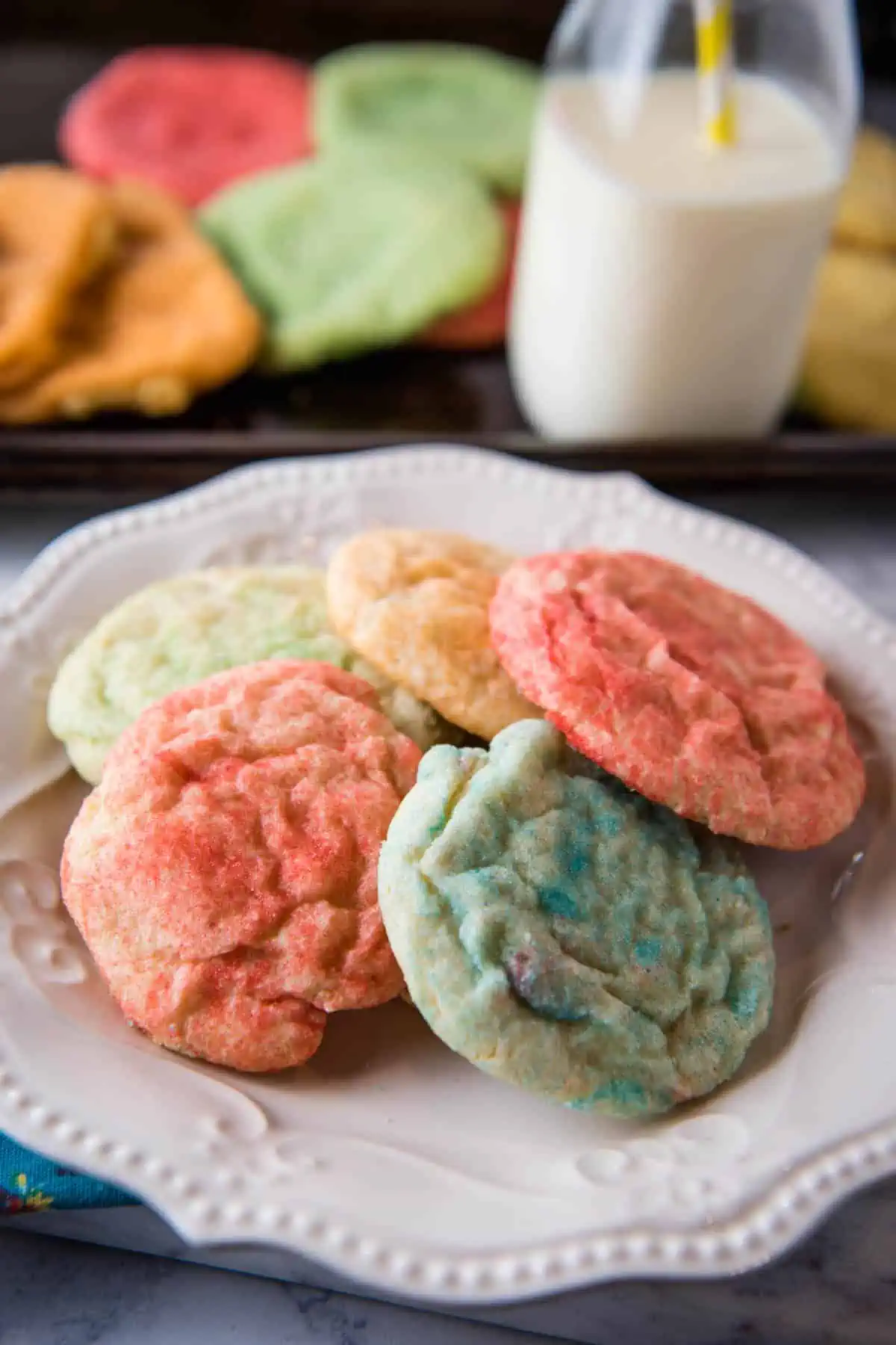 pile of colorful Jello cookies on white plate with glass of milk and more cookies on baking sheet