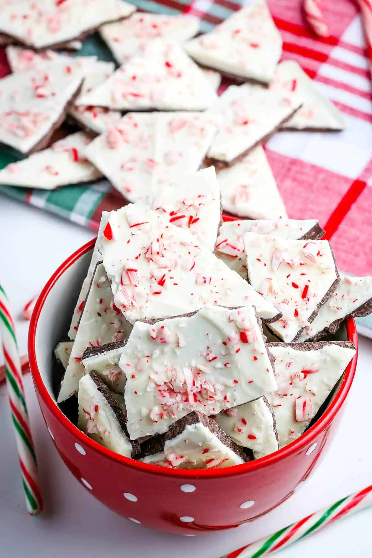 pile of white chocolate peppermint bark in red bowl and on plaid Christmas towel on white countertop with candy canes