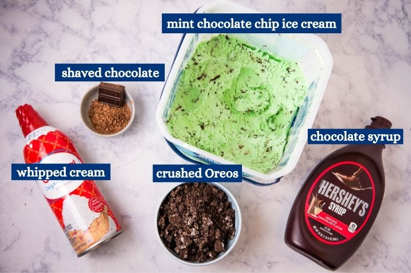 ingredients for grasshopper sundae, including shaved chocolate, mint chocolate chip ice cream, whipped cream, crushed Oreos, and chocolate syrup, on white marble countertop