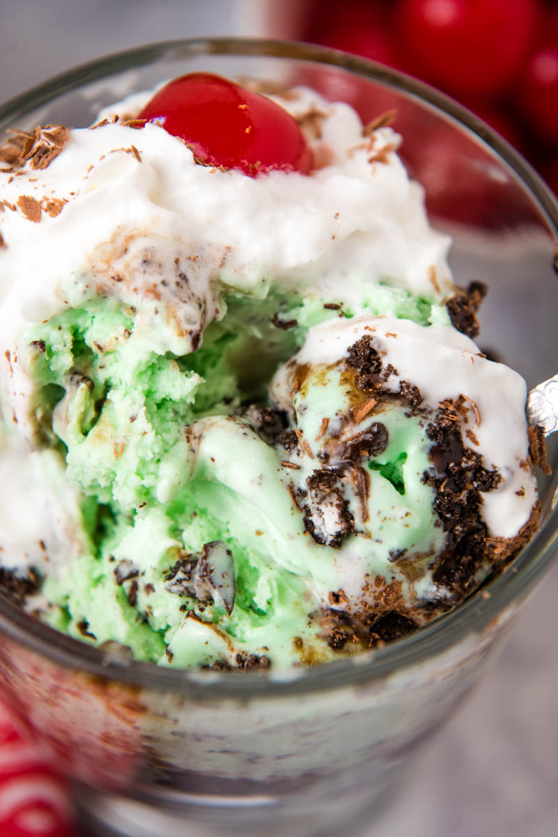 spoon digging into mint chocolate chip ice cream sundae with whipped cream and cherry on top, in clear glass sundae bowl