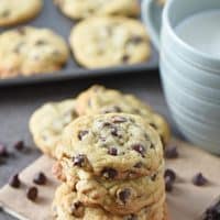 chocolate chip cookies stacked on brown paper bag with chocolate chips and blue mug of milk