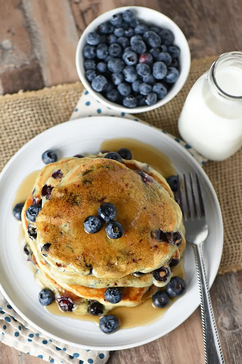 Blueberry Buttermilk Pancakes with maple syrup on a plate, fresh blueberries in a bowl, and milk in a glass bottle