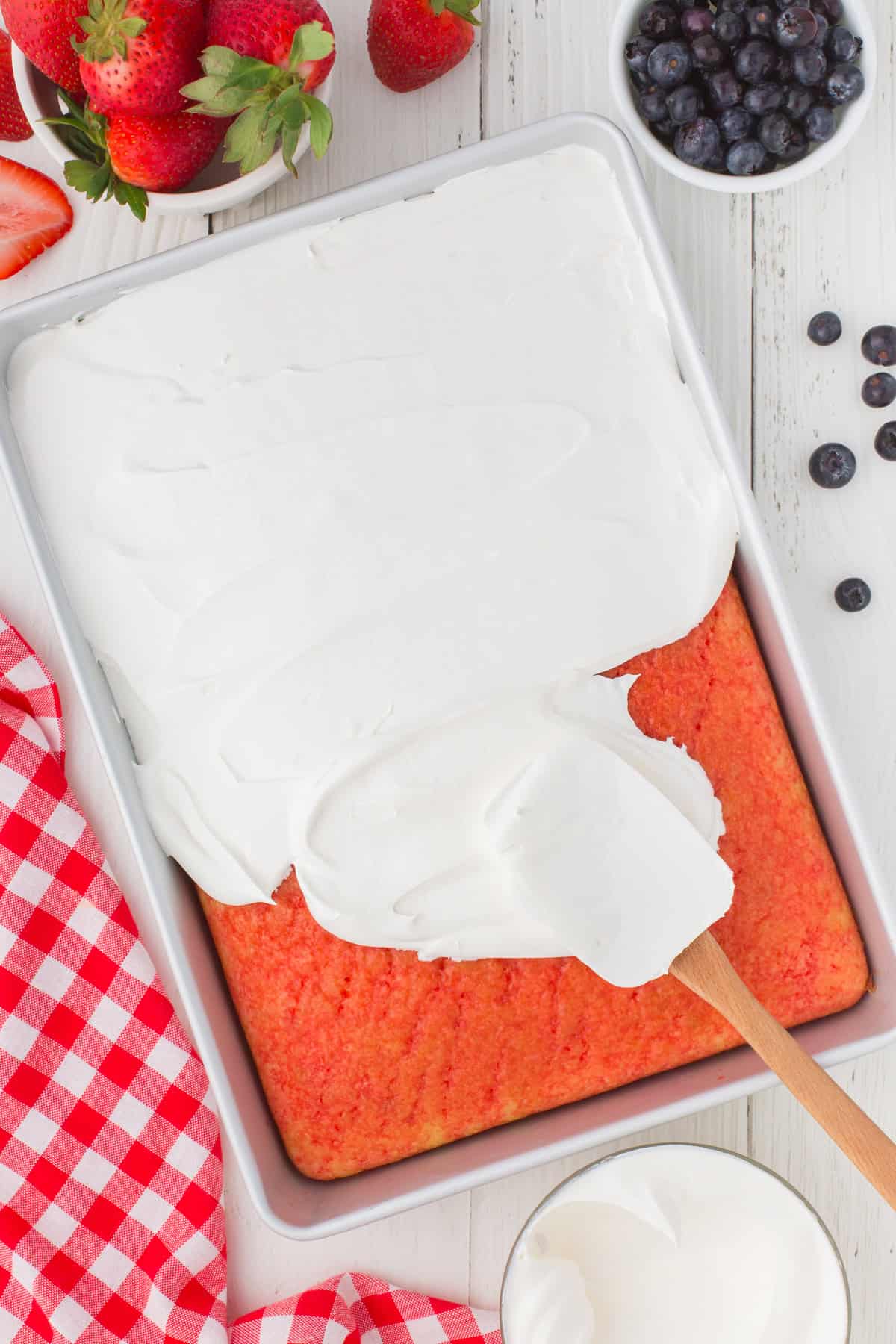overhead shot showing a layer of fluffy whipped topping being spread over the top of a strawberry Jello poke cake in a 9x13 cake pan.