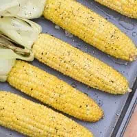 grilled corn in the husk on a cookie sheet with husks removed, seasoned with honey butter, salt, and pepper
