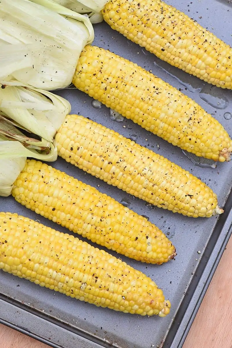 grilled corn in the husk on a cookie sheet with husks removed, seasoned with honey butter, salt, and pepper