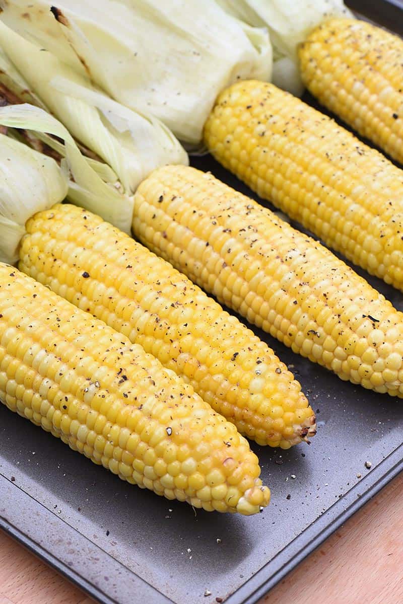 grilled corn on the cob on a cookie sheet with husks pulled back, seasoned with honey butter, salt, and pepper