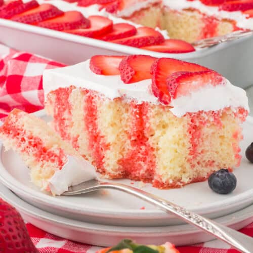 slice of strawberry Jello American flag cake on a small gray plate with the full 4th of July cake with berries in the background