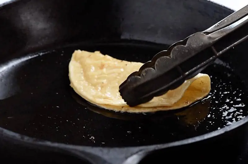 frying a corn tortilla into a taco shell in a cast iron skillet with tongs