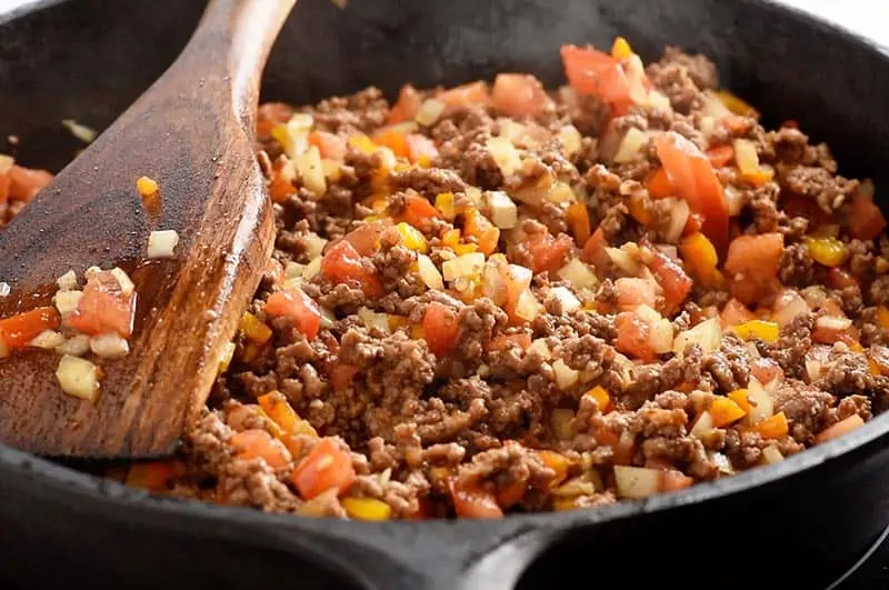 cooking ground beef with onion, peppers, and tomatoes in a cast iron skillet for a delicious taco recipe