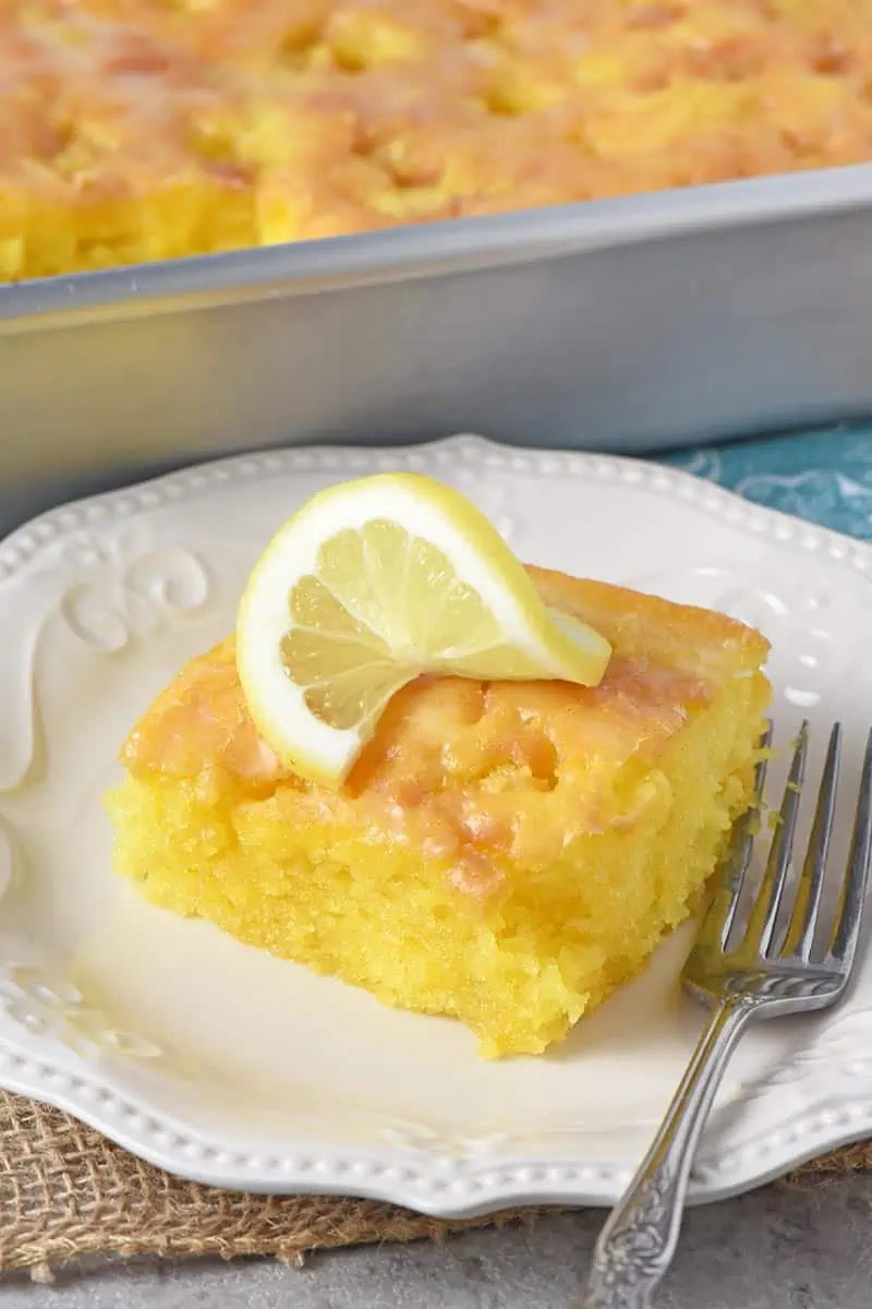 slice of lemon poke cake with lemon glaze on white plate with slice of lemon on top