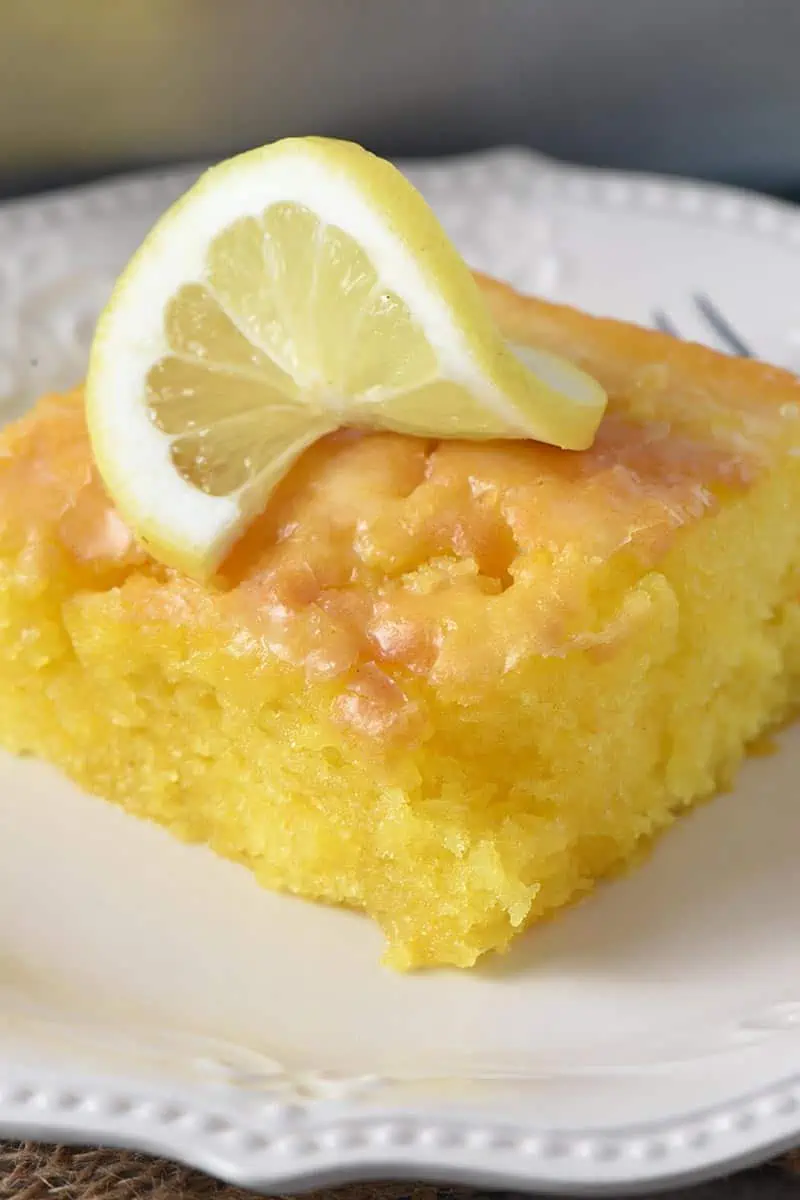 close up slice of lemon pudding cake with slightly crispy lemon glaze and slice of lemon on white plate