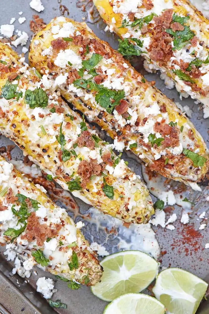 Mexican corn on a baking sheet with mayo, feta cheese, cilantro, bacon, and lime wedges