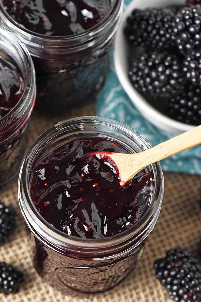 close up jar of homemade blackberry jam with tiny wooden spoon and fresh blackberries