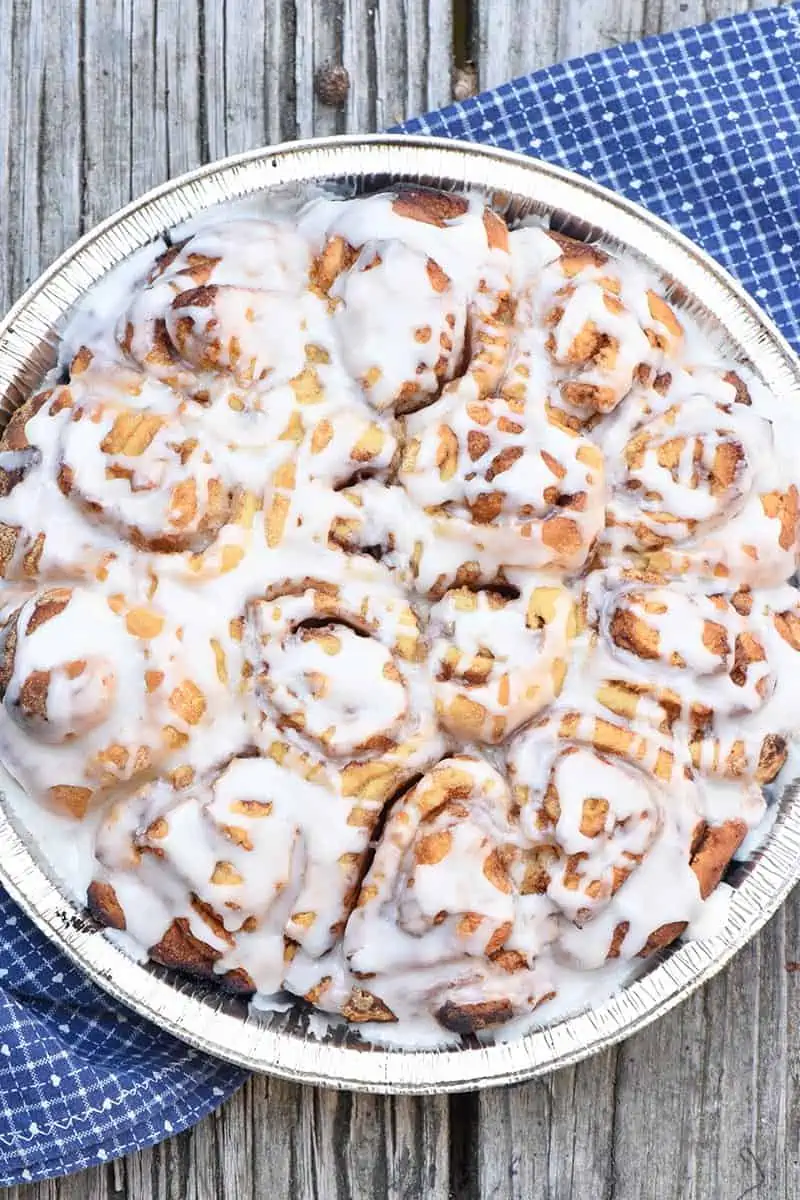 frosted camping cinnamon rolls in a foil pie plate on a wooden surface with a blue cloth