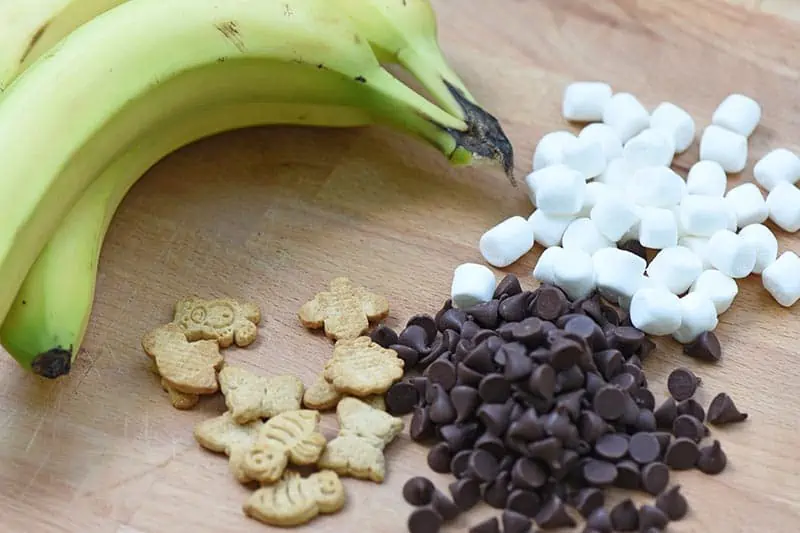 ingredients for banana boats s'mores on a cutting board, including chocolate chips, mini marshmallows, Teddy Grahams, and bananas