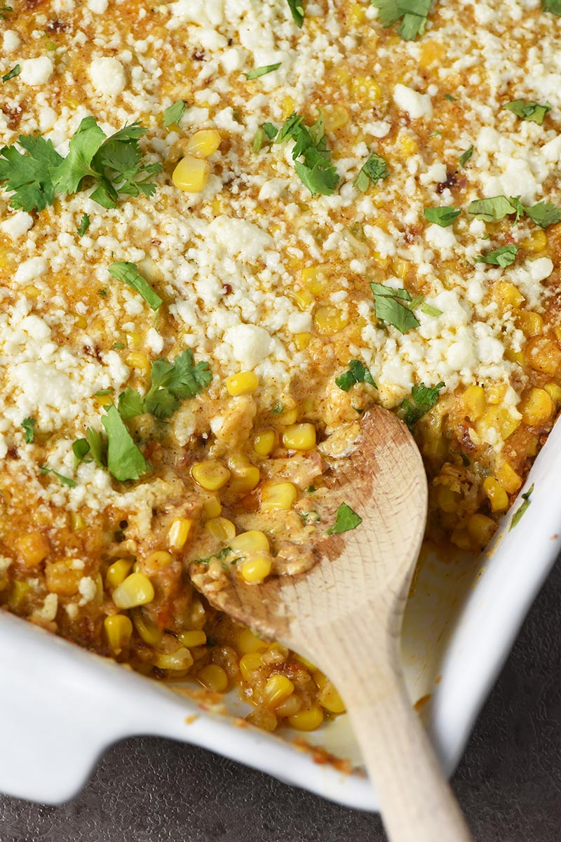 serving Mexican corn casserole with a wooden spoon out of a white baking pan