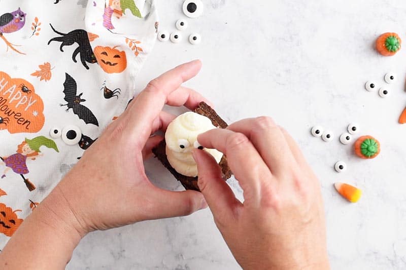 adding candy eyeballs to whipped cream ghost halloween brownies for easy halloween treats