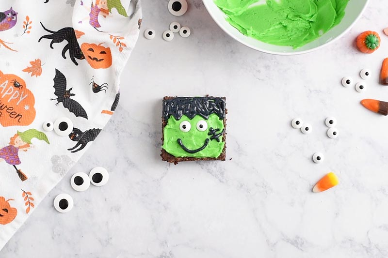 Frankenstein brownie on white marble countertop with green frosting, Halloween kitchen towel, candy eyeballs, and candy corn