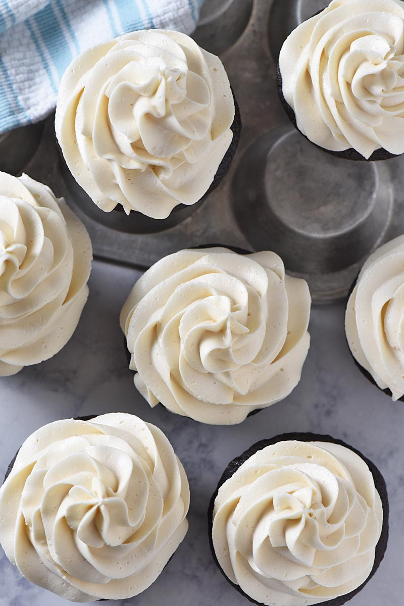 easy butterbeer buttercream frosting swirled onto chocolate cupcakes on white marble countertop with muffin tin and light blue kitchen towel