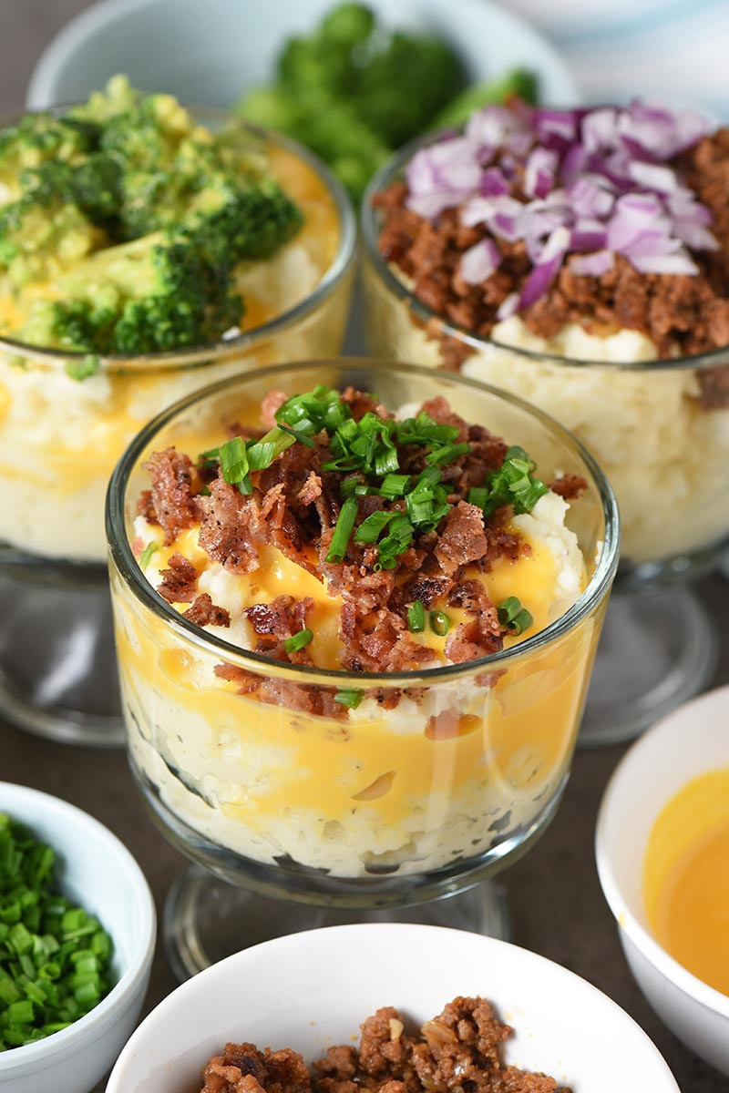 mash potato bar with mini trifle bowls of mashed potatoes covered in toppings like bacon, cheese sauce, chives, broccoli, sloppy joe meat, and red onion