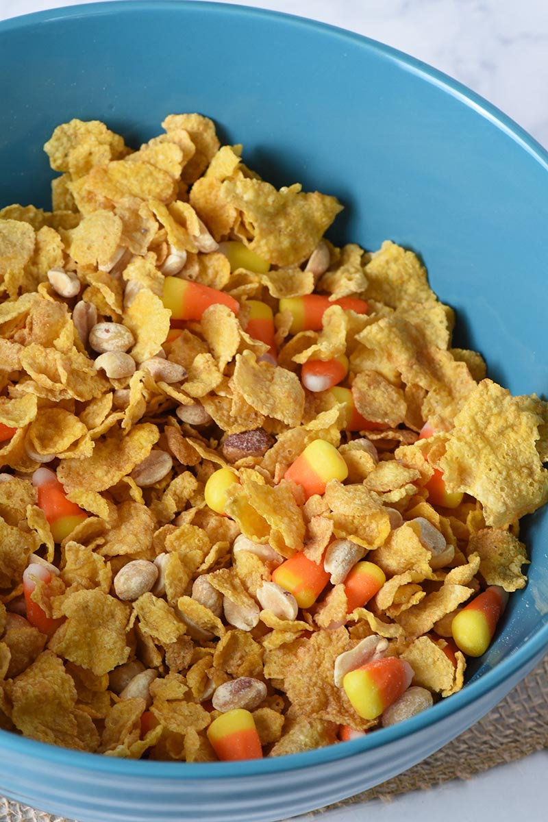 mixing together no bake peanut butter bars ingredients, including corn flake cereal, PLANTERS peanuts, and candy corn in large blue mixing bowl