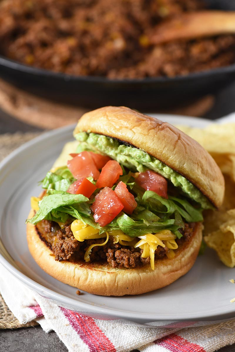 open faced taco sloppy joes sandwich with avocado, chopped tomatoes, lettuce, and shredded cheddar on a gray plate with tortilla chips