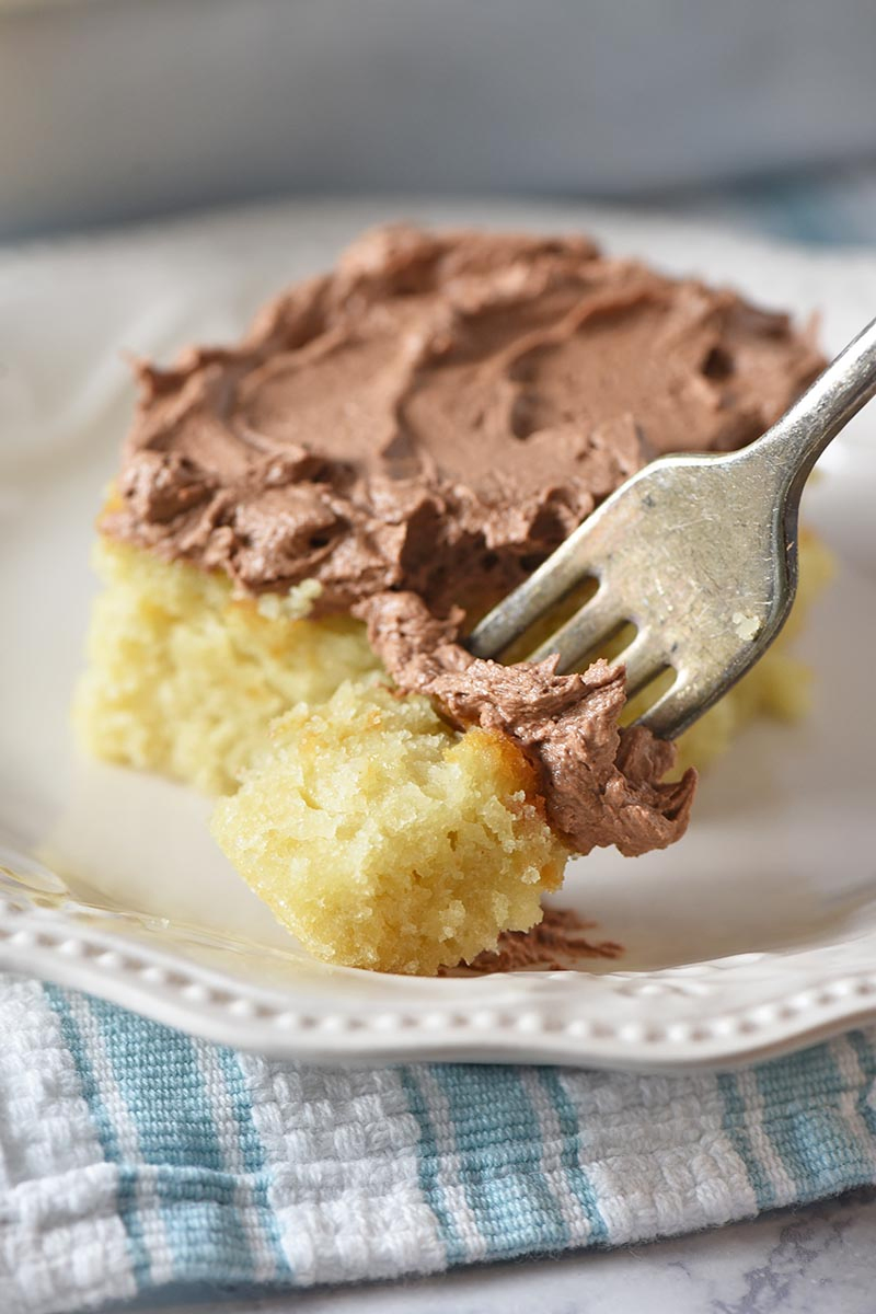 bite of yellow cake with chocolate frosting on a fork with ivory plate and blue and white striped kitchen towel