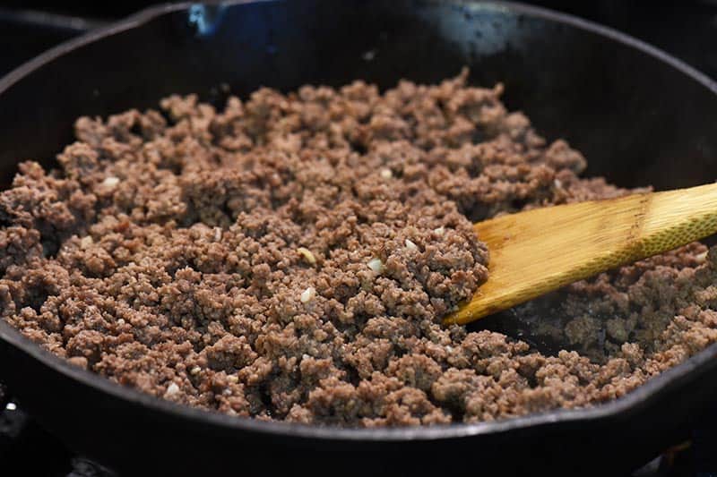 mixing ground beef and garlic together for pizza sloppy joe recipe in iron skillet with wooden spatula