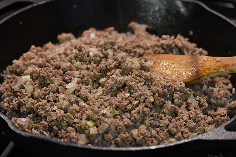 cooking ground beef, onion, and garlic for sloppy joe stuffed peppers recipe in cast iron skillet on stovetop