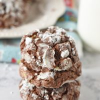stack of chocolate crinkle cookies on white countertop with plate of Christmas cookies and milk