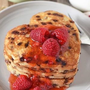 heart shaped pancakes with chocolate chips stacked on gray plate, slathered with raspberries and strawberry jam