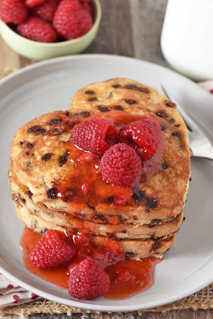 heart shaped pancakes with chocolate chips stacked on gray plate, slathered with raspberries and strawberry jam