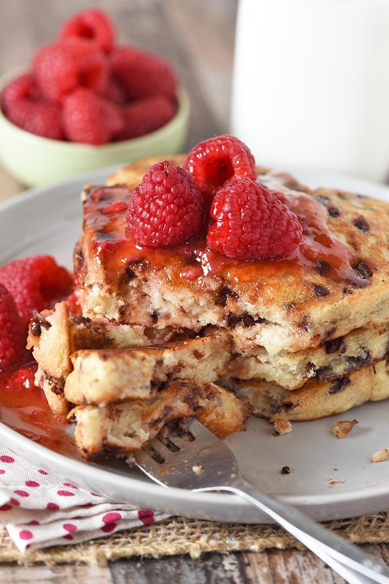 fork and bite of heart shaped chocolate chip pancakes with raspberries and sauce on gray plate