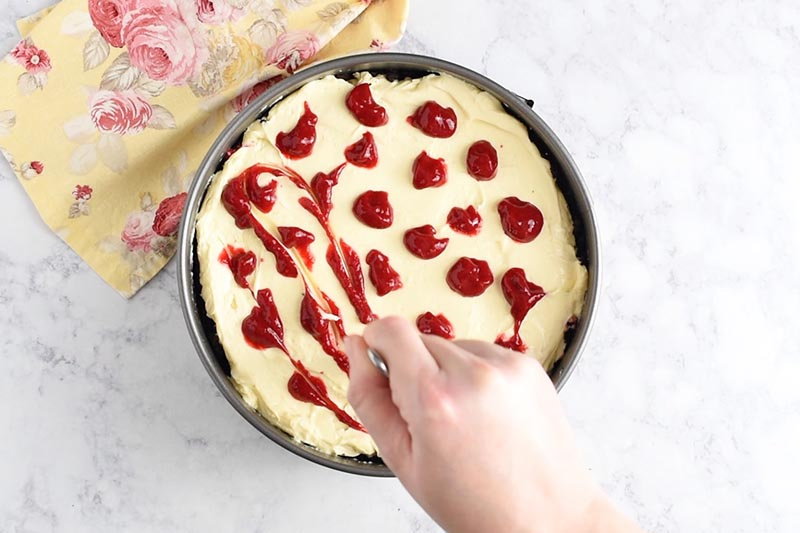 making raspberry swirl cheesecake by swirling raspberry sauce into cheesecake batter with a table knife