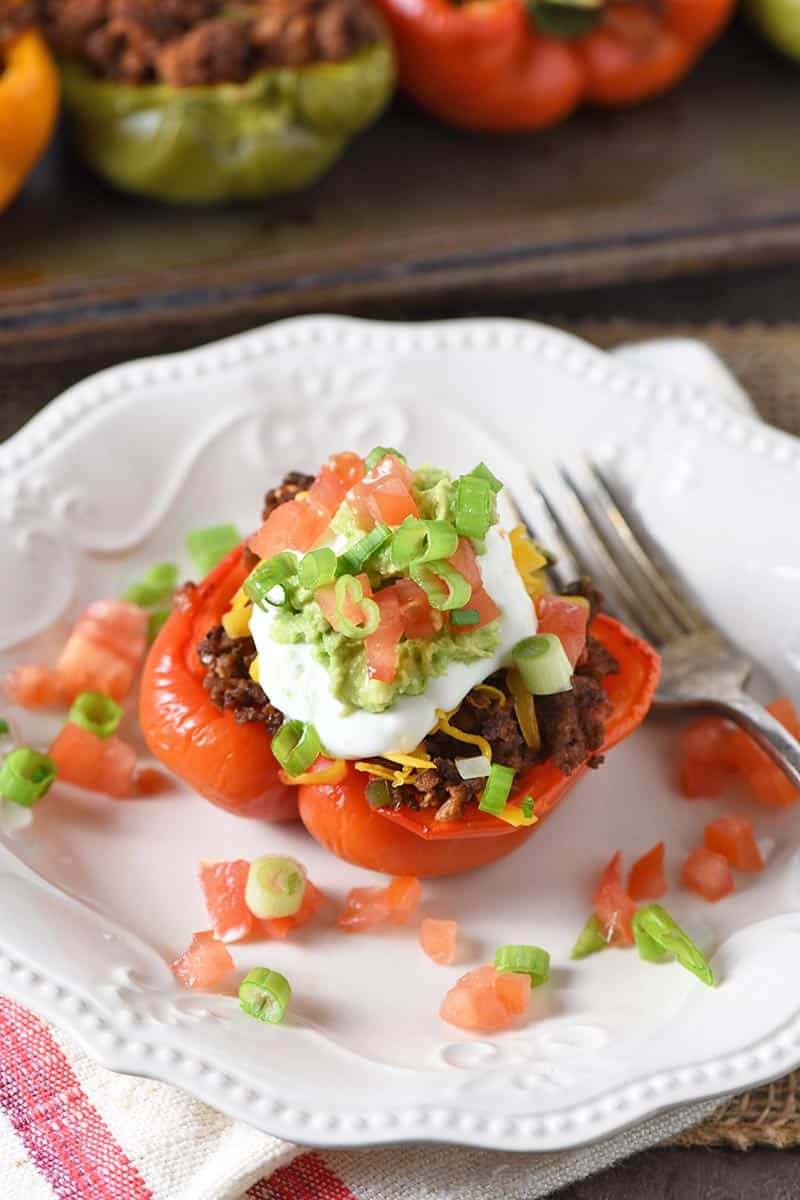 Mexican stuffed peppers, red pepper filled with ground beef taco meat and topped with taco toppings like lettuce, avocado, cheese, tomatoes, sour cream, and green onions, on a white plate