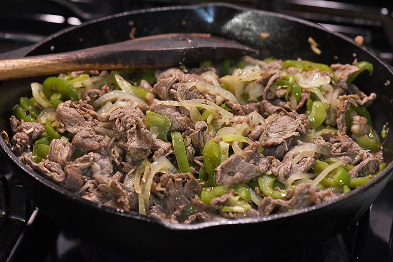 cooking Philly cheese steak meat with vegetables in cast iron skillet with wooden spatula