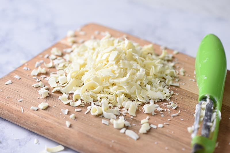 shredding white chocolate with a vegetable peeler on a wooden cutting board