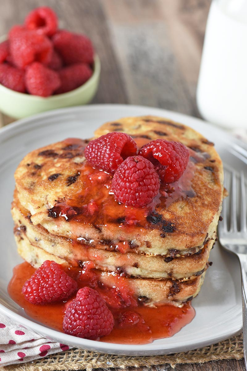 stacked chocolate chip Valentine pancakes in heart shapes for Valentine's Day, Mother's Day, and more on a gray plate with raspberries and syrup
