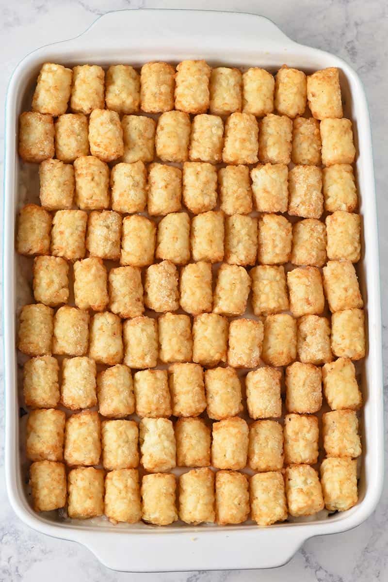 layering tater tots on tater tot hotdish for baking, in blue and white casserole dish