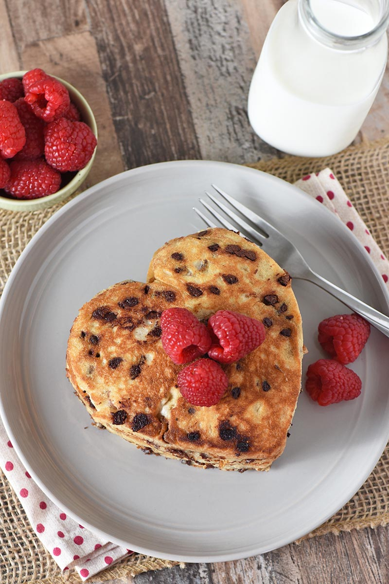 pancake recipe, heart shaped chocolate chip pancakes on gray plate with raspberries, sauce, fork, and milk