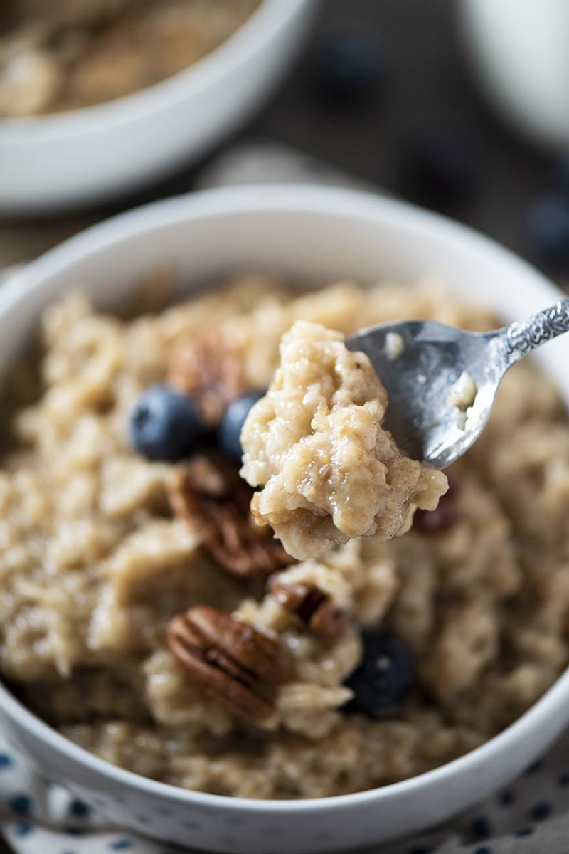 spoonful of maple and brown sugar oatmeal above white bowl of oatmeal with pecans and blueberries