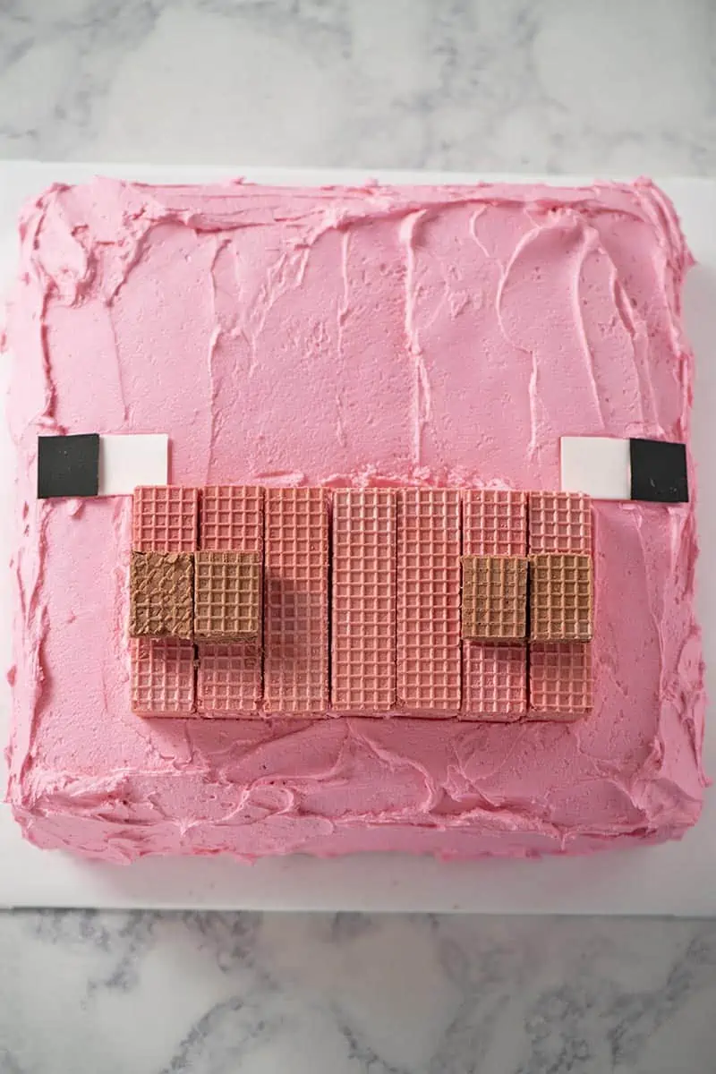 Minecraft pig cake decorated with sugar wafer cookies, sugar paper, and pink buttercream frosting on white cake board.