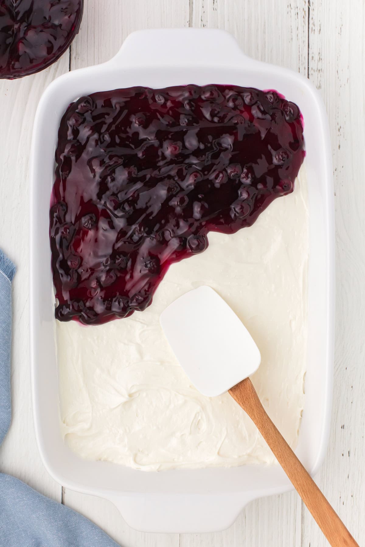 spreading the blueberry pie filling layer on top of the cream cheese layer for blueberry delight in a white casserole pan