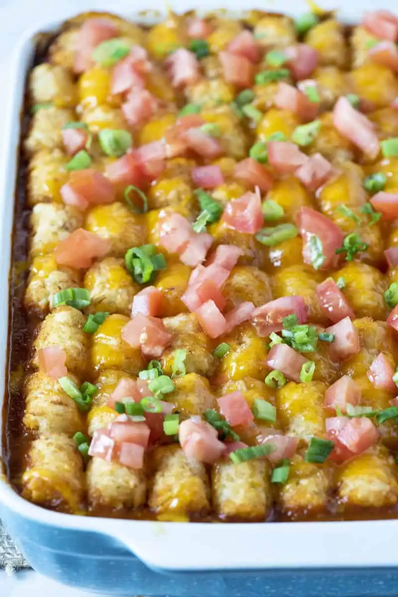 taco tater tot casserole topped with tomatoes and green onions in a blue baking dish