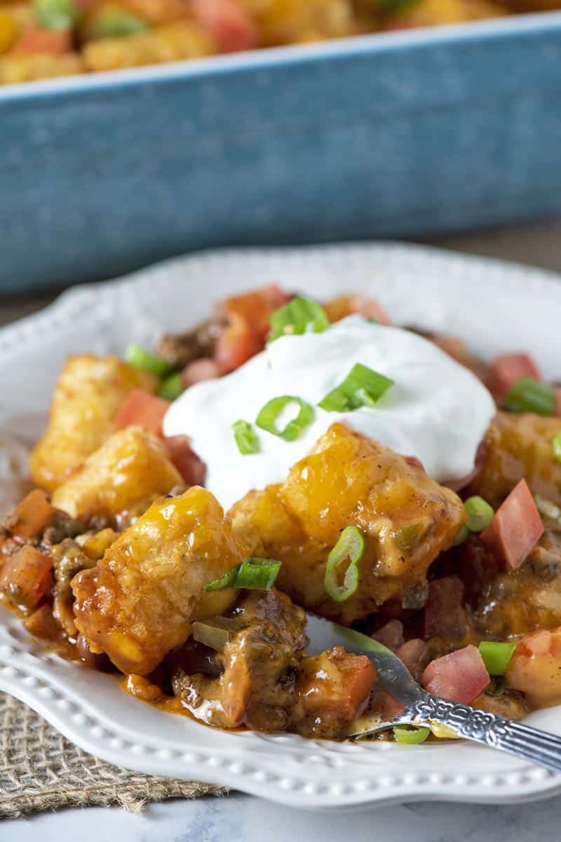 Mexican tater tot casserole on white plate with fork, also topped with sour cream and green onions