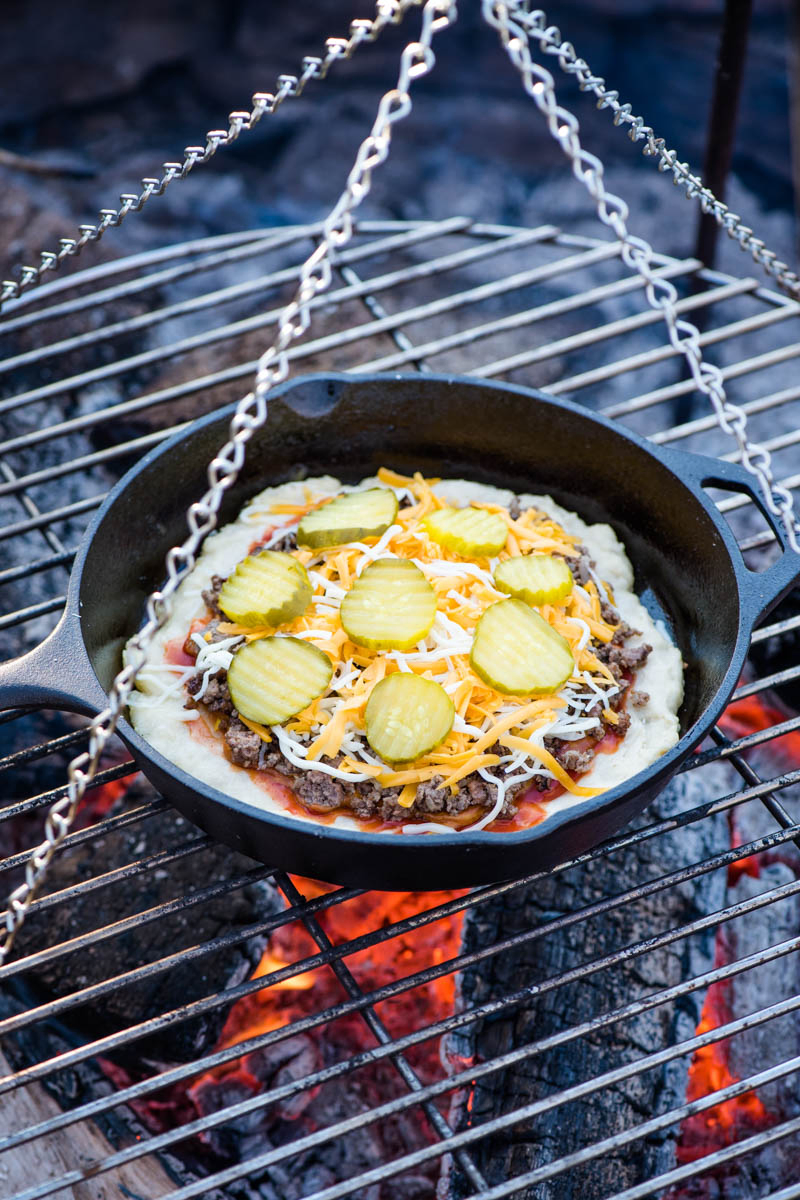 cooking hamburger pizza on a campfire in a cast iron skillet, using a tripod grill