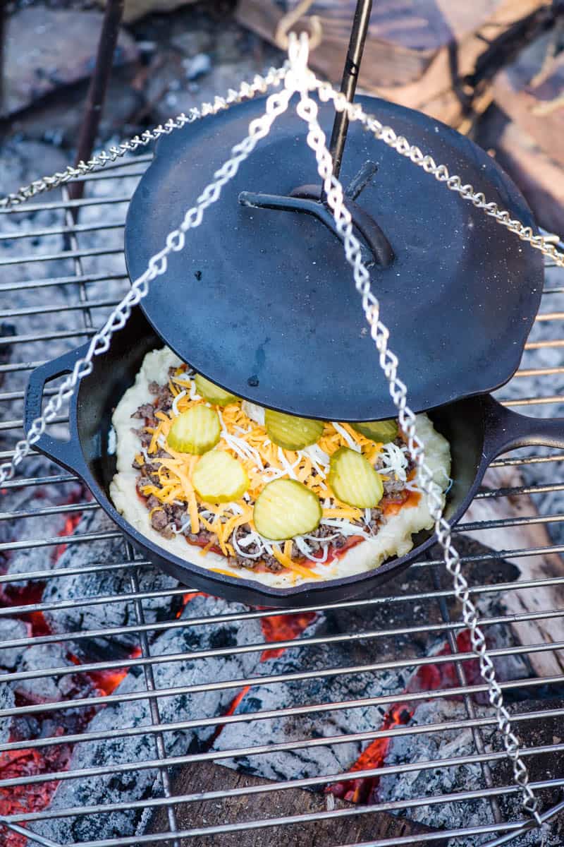 placing heated cast iron lid on cast iron pan for baking cast iron pizza on a campfire