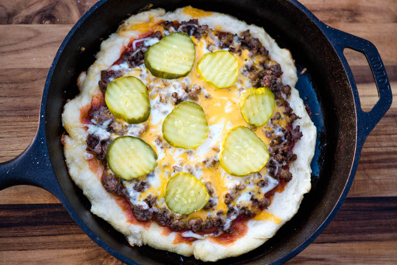 cooked or grilled cheeseburger pizza with pickles in cast iron skillet on wooden cutting board